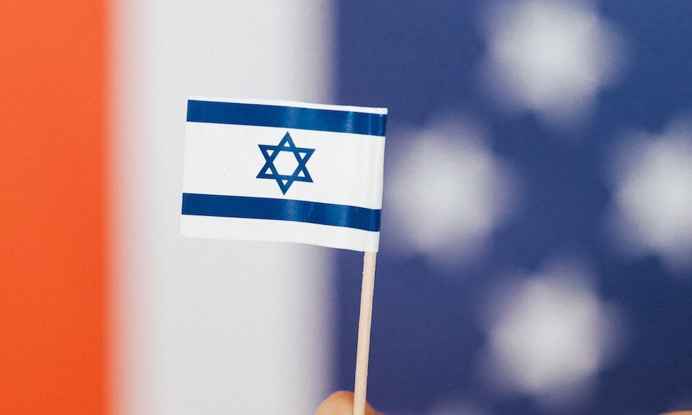 Someone holding a mini Israeli flag in front of the star spangled banner.